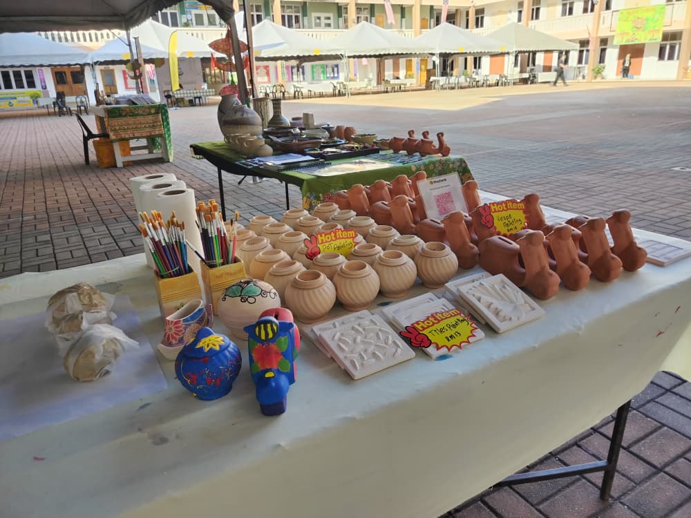 Rows of unpainted bisque pots and Labu Sayong forms beside jars of brushes and brightly painted finished examples